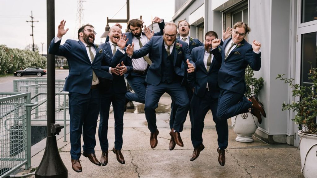 Groomsmen in blue suits jumping and celebrating outside Press Street Station in New Orleans